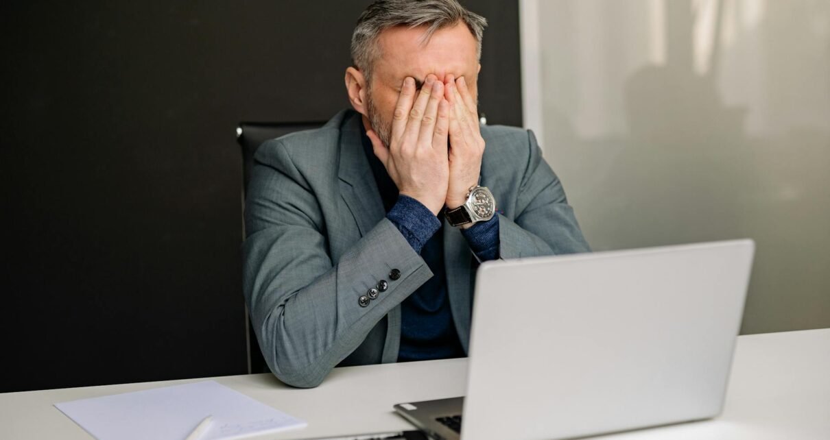 man in gray suit sitting on black chair covering his face